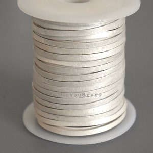 May include: A spool of silver-colored flat cord. The cord is tightly wound around a white plastic spool. The cord appears to be made of a soft, flexible material. The text "MichouBeads" is visible on the spool.