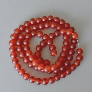 May include: A strand of frosted, round beads in a warm, reddish-orange hue. The beads are strung together, creating a circular pattern against a neutral gray background. The beads appear to be approximately 0.5 inches in diameter, suitable for jewelry making.