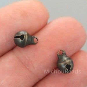 May include: Two small, antique bronze-colored bell charms. Each bell has a rounded body with a slit opening and a small loop at the top for stringing. The bells are likely for jewelry making or other crafts.