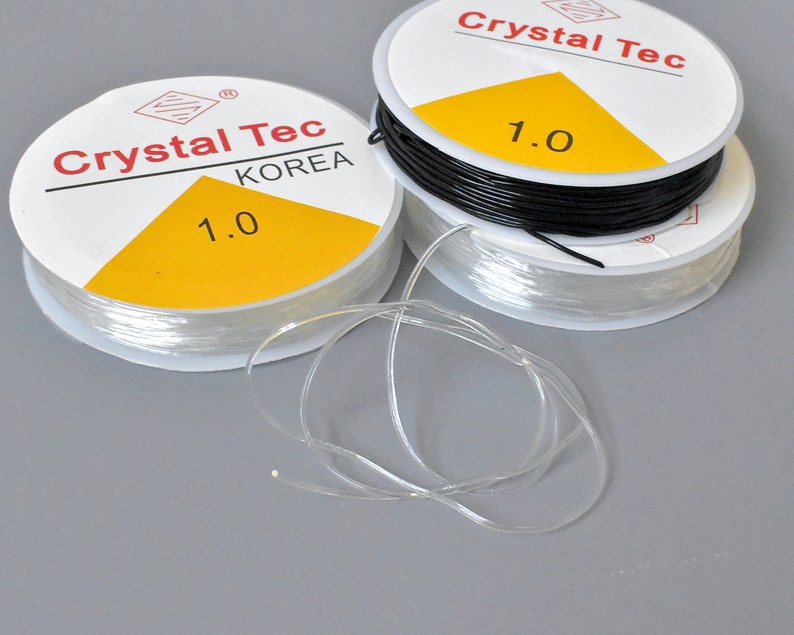 May include: Spools of clear and black elastic string, labeled "Crystal Tec KOREA 1.0". The clear string is unwound, showing its flexibility. The spools have a white label with red text and a yellow triangle.