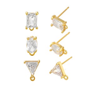 May include: A set of six gold-toned stud earrings featuring clear, faceted stones in rectangular, oval, and triangular shapes. Each earring includes a small metal loop at the base for attaching charms, with a post back for secure fastening.
