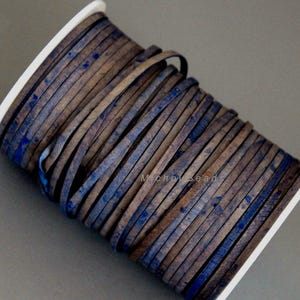 May include: A spool of flat leather cord in shades of brown and blue. The cord is tightly wound on a white plastic spool. The leather has a distressed, mottled appearance, with streaks and spots of blue against a brown base.