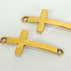 May include: Two gold-colored cross charms with a loop at each end. The crosses have a classic design with a slightly curved bar. The charms are likely for jewelry making or crafting.