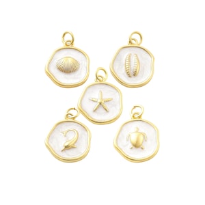 May include: Five gold-colored pendants with white enamel backgrounds. Each pendant features a different sea-themed design: a seashell, a starfish, a dolphin, a turtle, and a pair of seashells. The pendants have a gold-colored border and a loop for attaching to a chain.