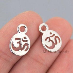May include: Two silver-colored Om symbol charms. Each charm has an oval shape with the Om symbol cut out in the center. The charms have a small loop at the top for attaching to jewelry. The charms are shown against a blurred background.