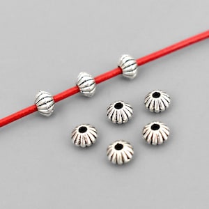 May include: Silver-colored metal beads in two distinct designs. Some are barrel-shaped with vertical grooves, strung on a red cord. Others are flower-shaped with a central hole. The beads are scattered on a gray surface, ideal for jewelry making.