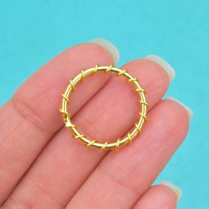 May include: A gold-colored ring with a wrapped design. The ring is held in a hand against a turquoise background. The ring's design features a series of gold-colored wraps around the band, creating a textured appearance.