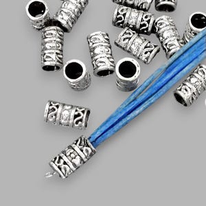 May include: Silver-colored metal beads with an intricate design, scattered on a gray surface. A blue cord is visible, threaded through a bead. The beads are cylindrical with a large hole. Ideal for jewelry making.