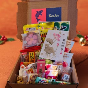 Puede incluir: Una caja de cartón llena de varios bocadillos y golosinas asiáticas. La caja contiene caramelos envasados, frutas secas y una tarjeta con una ilustración de flor de loto. Una tarjeta roja y azul con un gráfico de pez y el texto "KoiJoy" es visible.