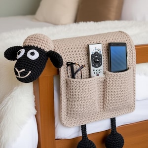 May include: A beige crochet bed organizer shaped like a sheep, with a black face and ears. It has pockets for a remote, phone, and glasses. The sheep's head is on the left, and two black pom-poms hang below.