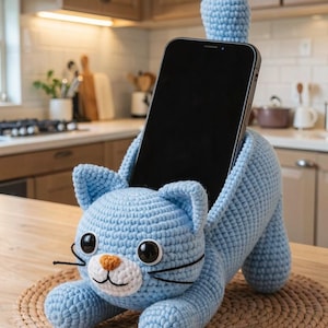 以下が含まれることがあります： 水色の編み猫のスマホホルダー。黒いスマートフォンがセットされています。猫は大きな黒い目、白い口、オレンジ色の鼻をしています。ホルダーは木製の表面にある織りマットの上に置かれています。