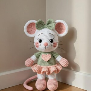 K&ouml;nnte beinhalten: Handgefertigte H&auml;kelmaus-Puppe mit wei&szlig;em K&ouml;rper, rosa Ohren und gr&uuml;ner Schleife. Die Maus tr&auml;gt ein gr&uuml;nes Oberteil mit rosa Herz und einen rosa R&uuml;schenrock. Die Puppe hat schwarze Knopfaugen und lange Schnurrhaare.