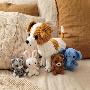 May include: A collection of handmade crochet stuffed animals, including a large dog with brown and white markings, a gray cat, a white bunny, a brown bear, and a blue elephant. The toys are arranged on a bed with neutral-toned pillows.