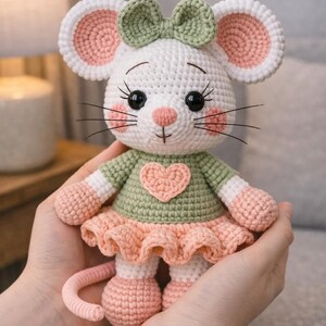 K&ouml;nnte beinhalten: Eine handgefertigte H&auml;kelmauspuppe mit wei&szlig;em K&ouml;rper, rosa und wei&szlig;en Ohren und einer gr&uuml;nen Schleife. Die Maus tr&auml;gt ein gr&uuml;nes Oberteil mit einem rosa Herz und einen rosa Rock. Die Puppe wird in zwei H&auml;nden gehalten.