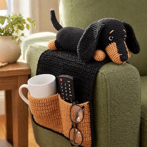 Pode incluir: Um cão dachshund de crochê preto e castanho repousa sobre um organizador de braço de sofá de crochê preto. O organizador tem bolsos para uma caneca branca, controle remoto e óculos. O sofá é verde.