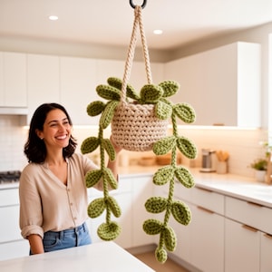 May include: A crocheted hanging plant with a beige pot and green leaves. The plant is suspended from a black hook by a beige rope. The leaves cascade downwards, creating a decorative and natural aesthetic. The plant is displayed in a kitchen setting.