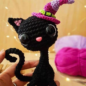 May include: A handmade black crochet cat with large black eyes, a pink nose, and a curly tail. The cat wears a witch hat in shades of pink, purple, and green. The cat is a whimsical, decorative item.