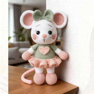 K&ouml;nnte beinhalten: Eine handgefertigte H&auml;kelmaus-Puppe mit wei&szlig;em K&ouml;rper, rosa Ohren und einer gr&uuml;nen Schleife. Die Maus tr&auml;gt ein gr&uuml;nes Hemd mit einem rosa Herz und einen rosa Rock. Die Puppe hat schwarze Augen und rosa Wangen.