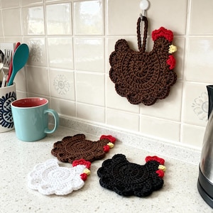 Puede incluir: Una colección de artículos hechos a mano con forma de pollo de ganchillo. La imagen muestra un salvamanteles de pollo marrón colgado de un gancho de pared y tres posavasos con forma de pollo en blanco, marrón y negro.