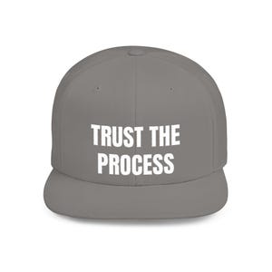 Peut inclure: Casquette de baseball grise avec visière plate, portant l'inscription blanche "TRUST THE PROCESS" en lettres grasses. Le chapeau a une couronne structurée et une visière plate.