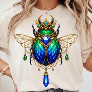 May include: A cream-colored t-shirt featuring a detailed illustration of a jeweled beetle. The beetle has a green and blue body, gold wings, and a crown. Dangling gems and gold chains add to the design.