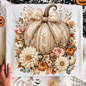 May include: A white t-shirt featuring a central graphic of a large, cream-colored pumpkin adorned with a decorative lace bow. The pumpkin is surrounded by a detailed arrangement of autumn flowers, smaller pumpkins, and foliage in shades of beige.