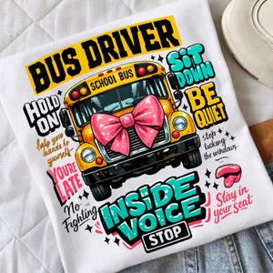 Può includere: T-shirt bianco con grafica di scuolabus giallo con fiocco rosa e testo che dice "BUS DRIVER", "HOLD ON", "INSIDE VOICE" e "SIT DOWN BE QUIET". La maglietta ha un design divertente e a cartoni animati.