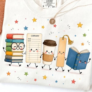 Pode incluir: Uma t-shirt cor de creme com um gráfico de objetos antropomórficos, incluindo uma pilha de livros coloridos com óculos, um cartão de biblioteca com o texto LIBRARY NAME DATE, um copo de café, um marcador de página e um livro azul aberto.