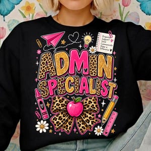 以下が含まれることがあります： ゴールドのヒョウ柄とピンクで「ADMIN SPECIALIST」と書かれた黒いスウェットシャツ。ピンクの紙飛行機、電球、テキスト付きのメモ帳、ピンクのクレヨン、ピンクの定規、鉛筆、リンゴが付いたヒョウ柄のリボンのグラフィックが描かれています。