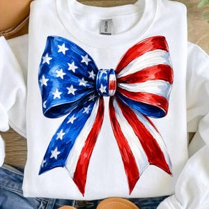 May include: A white crewneck sweatshirt featuring a large, centered graphic of a bow. The left side of the bow is blue with white stars, and the right side features red and white horizontal stripes, mimicking the American flag pattern.