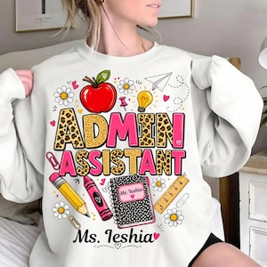 May include: A white crewneck sweatshirt featuring a graphic print with the text "ADMIN ASSISTANT" and "Ms. Ieshia". The design includes a red apple, yellow lightbulb, paper airplane, pencil, crayon, ruler, paperclips, and daisies.