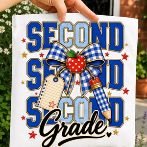 May include: A white tote bag featuring a graphic design with the text SECOND GRADE in blue block letters. The design includes a blue and white checkered bow, a red apple with white dots, a blank lined tag, a pencil, and scattered red and gold stars.
