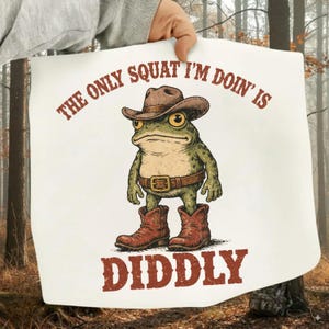 Pode incluir: Um cartaz branco com um sapo de desenho animado usando um chapéu de cowboy, cinto e botas. O texto diz "THE ONLY SQUAT I'M DOIN' IS DIDDLY" em letras marrons. O sapo é verde com olhos amarelos.