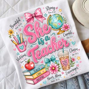 May include: White t-shirt featuring the text "She is a Teacher" in pink and teal. The design includes a pink bow, a globe, pencils, books, and a coffee cup. The shirt has inspirational words and Bible verses.