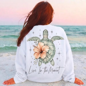Puede incluir: Una sudadera blanca de manga larga con un estampado gráfico en la espalda. El diseño muestra una tortuga marina con una gran flor de hibisco color melocotón, rodeada de pequeños iconos de estrellas y el texto Live In The Moment en negro.