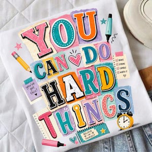 May include: White t-shirt with the words "YOU CAN DO HARD THINGS" in colorful, block letter designs. The shirt includes illustrations of a pencil, marker, and an alarm clock. Additional text includes "you've got this!" and "BELIEVE -IN- YOURSELF."