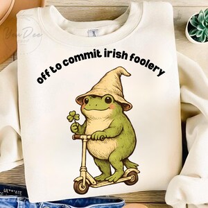 May include: Cream-colored sweatshirt with the text "off to commit irish foolery." The graphic features a green frog wearing a pointed hat and holding a four-leaf clover, riding a scooter. The design has a whimsical, cartoonish style.