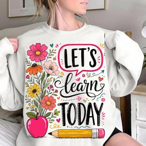 May include: A white crewneck sweatshirt featuring a colorful graphic print. The design includes a bouquet of pink, orange, and yellow flowers, a red apple, a yellow pencil, and the black text "LET'S learn TODAY" inside a pink speech bubble.