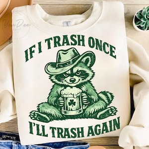 Könnte beinhalten: Cremefarbenes Sweatshirt mit einem grünen Aufdruck eines Waschbären mit Cowboyhut, der einen Bierkrug mit einem Kleeblatt hält. Der Text lautet "IF I TRASH ONCE I'LL TRASH AGAIN" in Grün. Ein lustiges, lässiges Oberteil.