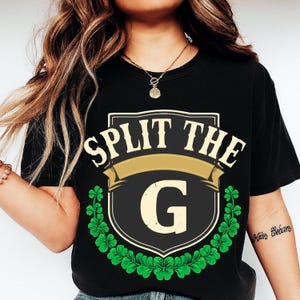 May include: Black t-shirt with a St. Patrick's Day design. The design features the text "SPLIT THE G" above a shield with the letter "G" and a gold banner, surrounded by green shamrocks.