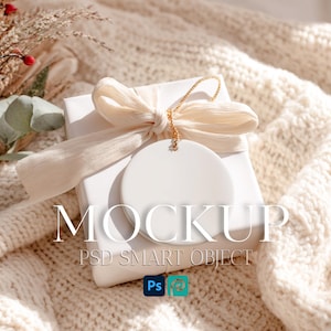 May include: A white square gift box topped with a cream-colored ribbon and a round white ceramic ornament hanging from a gold string. The box is placed on a textured cream knit fabric, with a sprig of dried eucalyptus and red berries nearby.