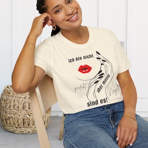 Könnte beinhalten: Cremefarbenes T-Shirt mit einer Grafik eines Frauengesichts, roten Lippen und schwarzer Nagelkunst. Der Text auf dem Shirt lautet "Ich bin nicht perfekt aber meine Nägel sind es!" in schwarzer Schrift. Das Model trägt eine blaue Jeans.
