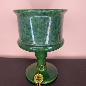 Può includere: Un vaso di ceramica verde con base a piedistallo. Il vaso ha un bordo ampio e svasato e una superficie strutturata. Un'etichetta dorata con la scritta "Prestige by Haeger" è attaccata alla base.