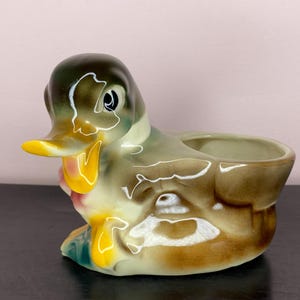 May include: A ceramic duck planter with a glossy finish. The duck has a yellow beak, a green head, and a brown body. The planter has a hollowed-out area for holding small plants or decorative items. The duck is sitting on a black surface.