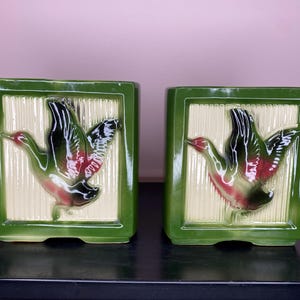 Peut inclure: Deux jardinières en céramique vertes de forme rectangulaire. Chaque jardinière présente un relief en forme de canard en vol, avec des couleurs noires, rouges et vertes. Le fond est de couleur crème clair avec des lignes verticales.