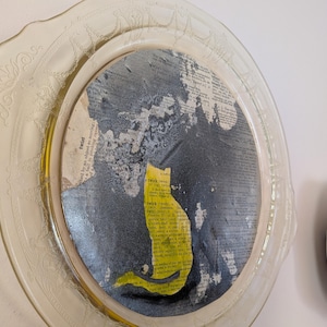 May include: A decorative plate with a yellow cat silhouette on a dark background. The cat is cut out of a yellow paper with text. The plate has a clear, ornate rim and is mounted on a wall.