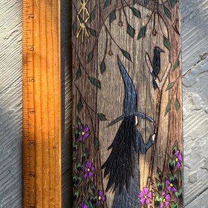 K&ouml;nnte beinhalten: Ein Holzbrandbild, das eine Hexe mit einem schwarzen Hut und langen schwarzen Haaren zeigt, die in einem Wald steht. Die Hexe h&auml;lt einen Stab, auf dem ein Rabe sitzt. Der Hintergrund zeigt einen Baum mit einem goldenen Runensymbol, gr&uuml;nen Bl&auml;ttern, violetten Blumen und roten Pilzen.