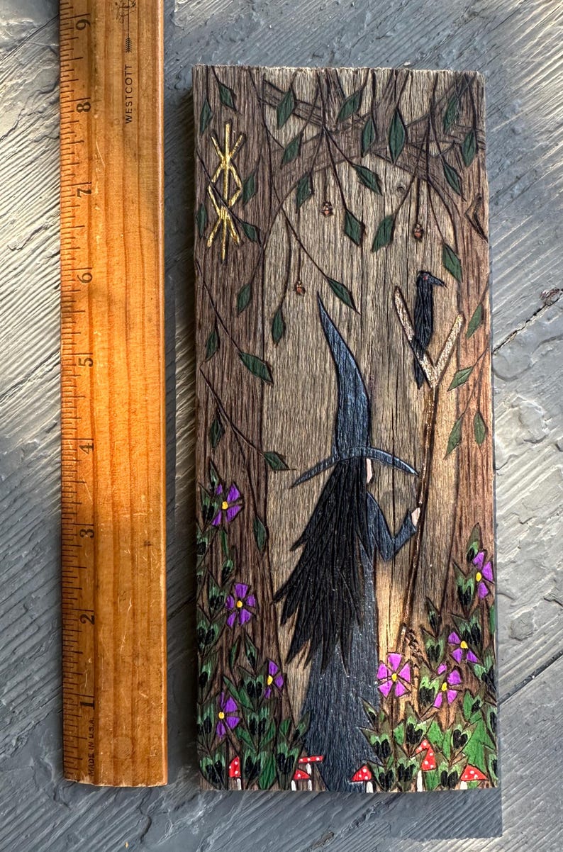K&ouml;nnte beinhalten: Ein Holzbrandbild, das eine Hexe mit einem schwarzen Hut und langen schwarzen Haaren zeigt, die in einem Wald steht. Die Hexe h&auml;lt einen Stab, auf dem ein Rabe sitzt. Der Hintergrund zeigt einen Baum mit einem goldenen Runensymbol, gr&uuml;nen Bl&auml;ttern, violetten Blumen und roten Pilzen.