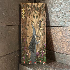 K&ouml;nnte beinhalten: Ein Holzbrandbild einer Hexe mit einem schwarzen Hut und langen schwarzen Haaren, die einen Stab mit einem darauf sitzenden Raben h&auml;lt. Die Hexe geht durch einen Wald mit lila Blumen und roten Pilzen.