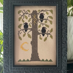 May include: A cross-stitch embroidery in a black ornate frame featuring a central brown tree with three black birds perched on its branches. A yellow crescent moon sits to the left, and a gold-colored geometric rune is stitched onto the tree trunk.